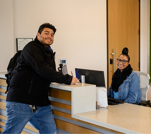 Smiling man leaning on a reception counter while a receptionist sits at her desk beside him.