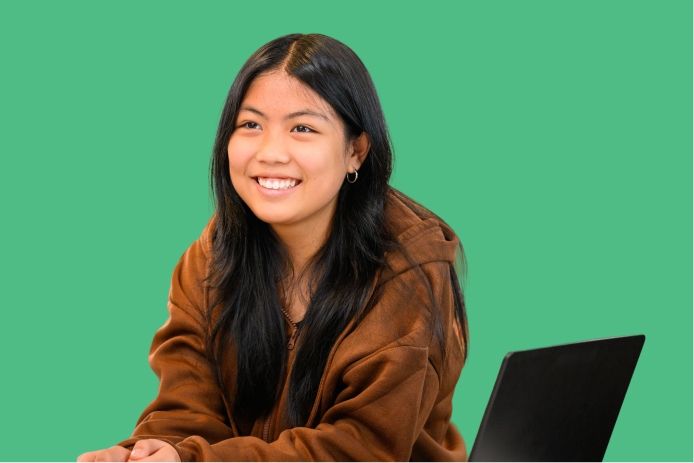Smiling young woman with long dark hair wearing a brown hoodie sits beside a laptop against a green background.